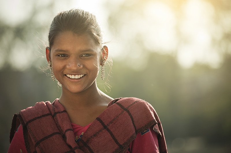 Outdoor image of a happy rural Indian girl giving toothy smile and looking at camera. She is in traditional dress which is Salwar Kameez and Dupatta.  One person, horizontal composition with selective focus.
